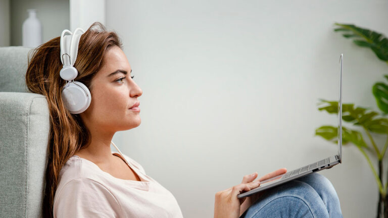 Nuori nainen kuuntelee äänikirjaa kotonaan valkoisista kuulokkeista läppäri sylissään.